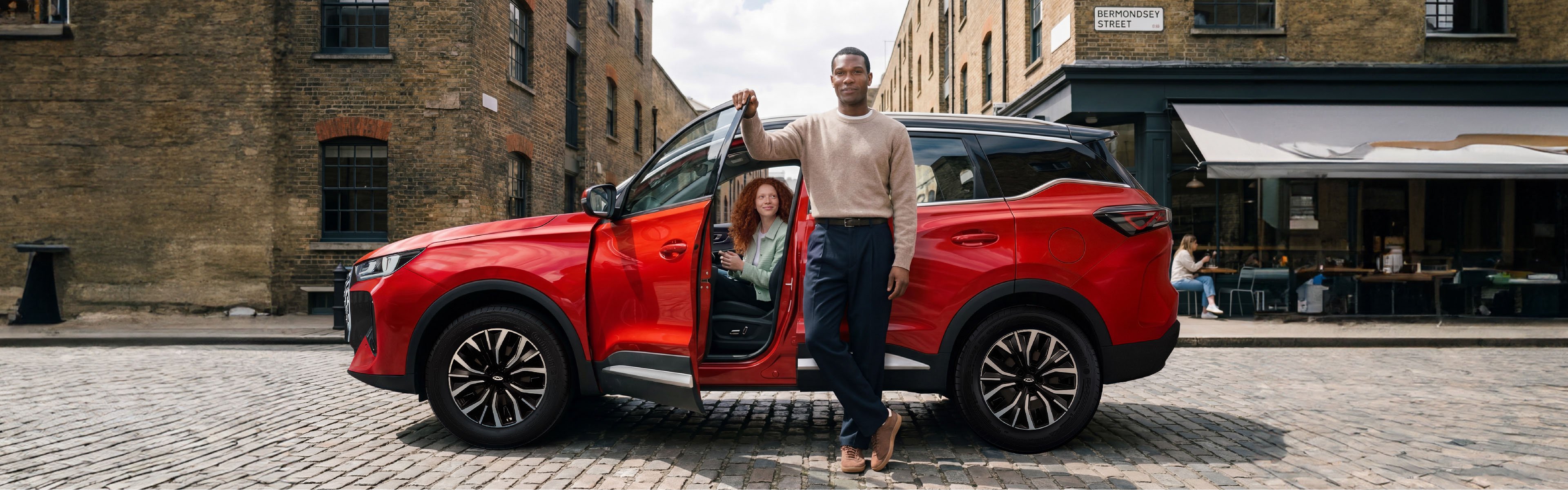 Man standing by an open door of a red SUV, with a woman seated inside.